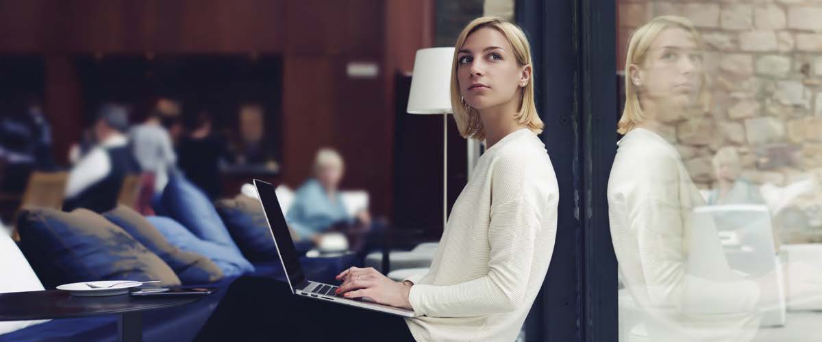 A young blonde woman leans against a glass window and looks up while operating a  laptop. Other students are blurry in the background, which appears to be a library or other study setting